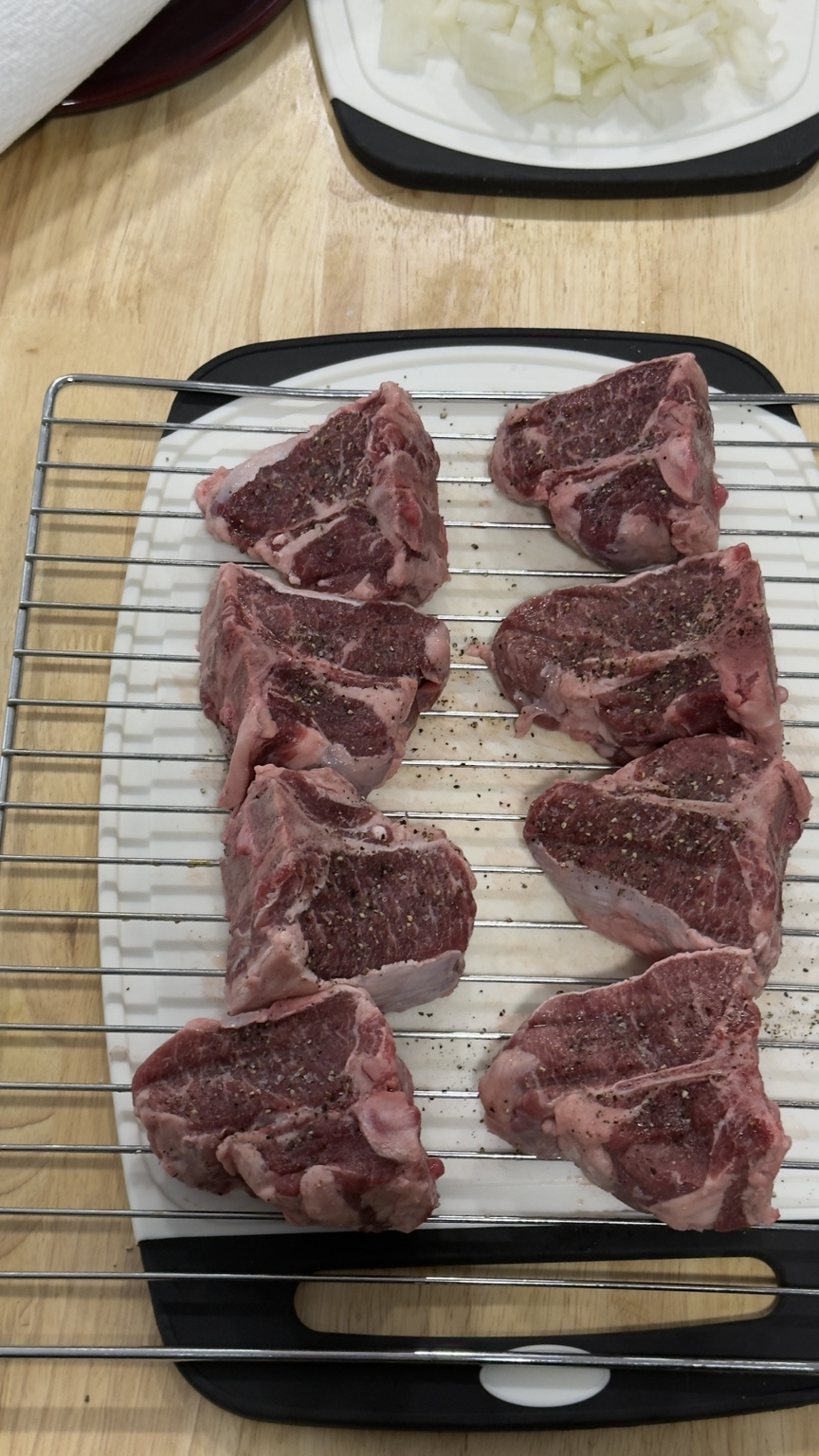 Raw bone-in lamb shoulder chops seasoned with kosher salt and black pepper on a wire rack.