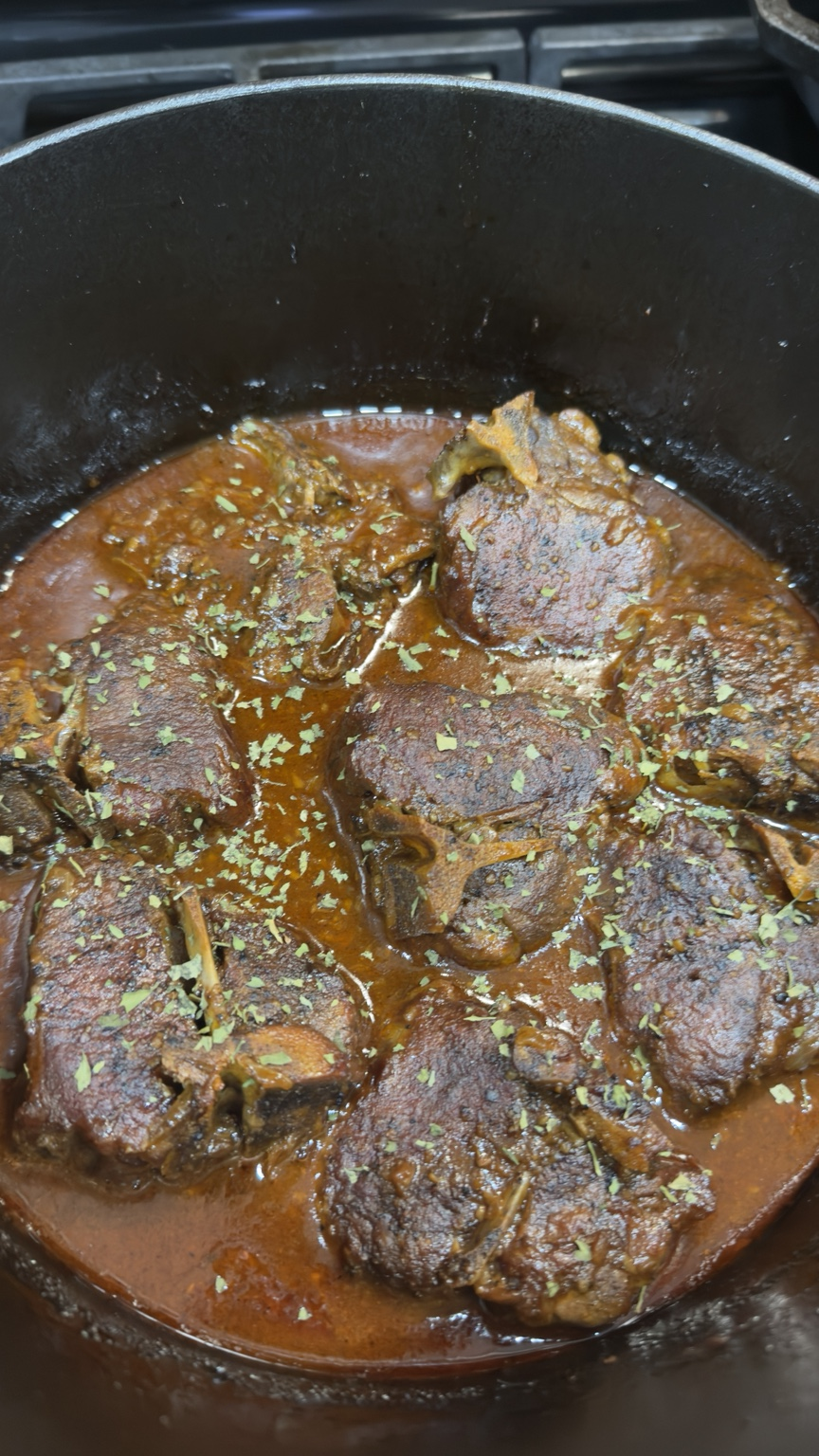 Braised lamb shoulder chops resting in a glossy reddish-brown onion sauce inside a black Dutch oven.